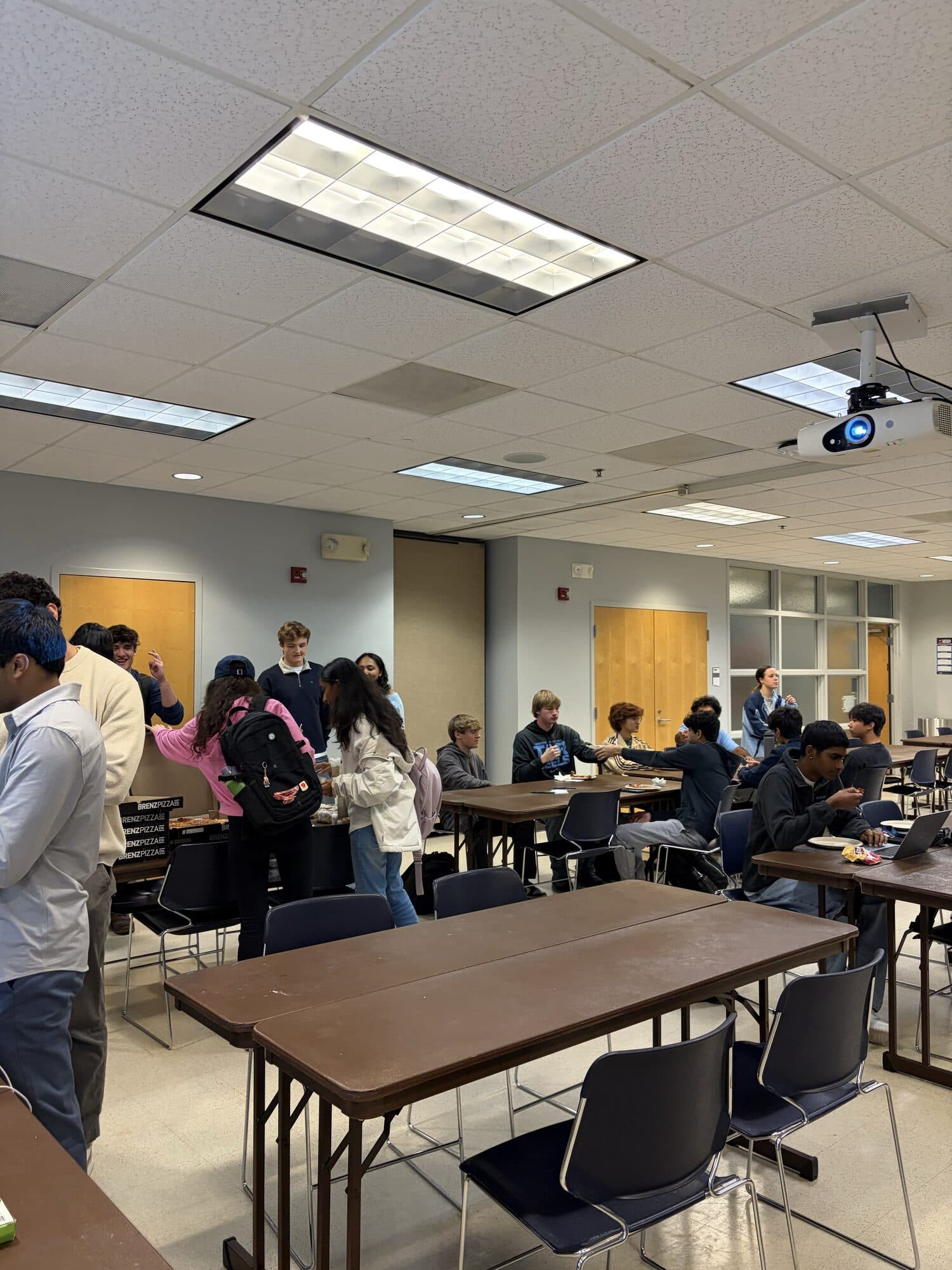 Trivia Night: students arriving and helping themselves to food at a side table while others sit at long tables under a ceiling projector.