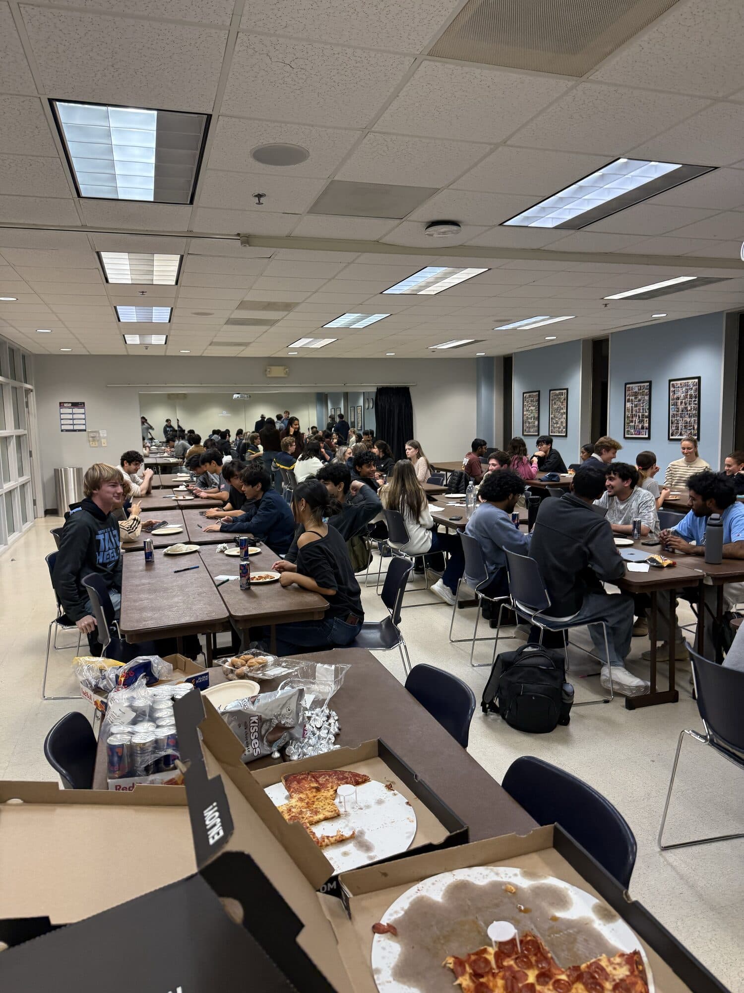 Trivia Night: pizza, drinks and snacks in the foreground and a full room of students talking at long tables.