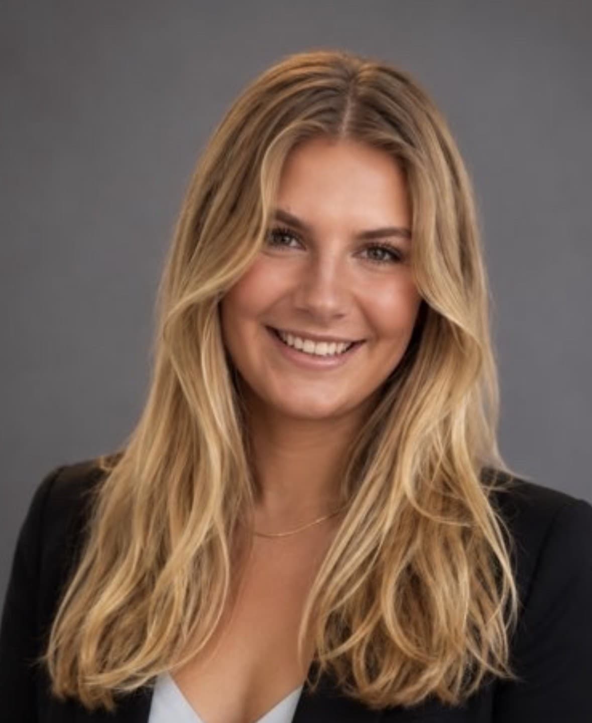 Harper Samples, head and shoulders, smiling at the camera in a dark blazer against a plain gray studio background.